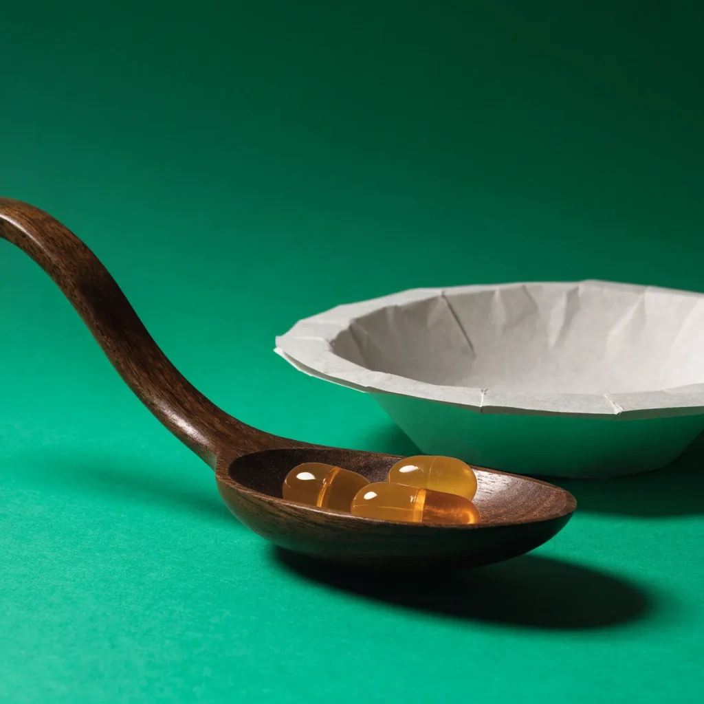 Three capsules in a wooden serving spoon beside a folded blank paper plate on a dark green surface
